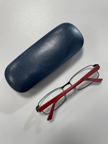 Görüntü, tahta bir masanın üzerinde duran bir güneş gözlüğünü gösteriyor. Güneş gözlükleri kırmızı çerçeveli siyahtır ve masa açık kahverengi renktedir. Güneş gözlüğü çerçevenin ortasına yerleştirilmiş ve arka planda hafif bir bulanıklık var.