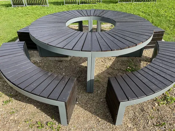A wooden picnic table with benches in a park. The table is set for a picnic, with a red checkered tablecloth, plates, cups, and food. There are two people sitting at the table, talking and laughing. The trees in the park are green and lush, and the s