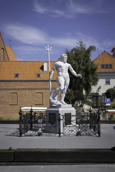 Mızrak tutan bir adamın heykeli kırmızı tuğla bir binanın önündedir. Heykel bir çit ve bir bankla çevrili.