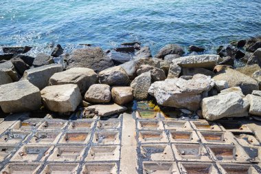 Arka planda büyük bir su kütlesi olan kayalık bir kıyı şeridi. Kıyı şeridi boyunca dağılmış kayalar karayla su arasında doğal bir bariyer oluşturuyor.