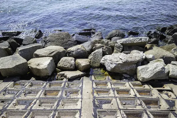 Arka planda büyük bir su kütlesi olan kayalık bir kıyı şeridi. Kıyı şeridi boyunca dağılmış kayalar karayla su arasında doğal bir bariyer oluşturuyor.
