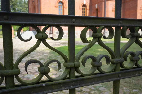 İşlenmiş demir bir çit, yeşil bir döşeme ve birkaç deliği var. Çit kırmızı çatılı bir binanın önünde.