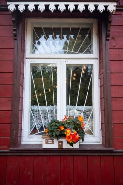 Beyaz perdeli bir pencere ve ev biçimli bir saksı. Pencere kırmızı bir binanın üzerinde.