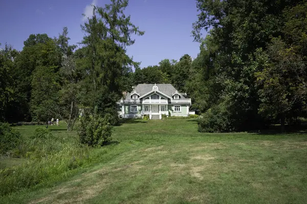 Yeşil çatılı büyük bir ev yemyeşil bir tarlada yer alır. Ev ağaçlarla çevrili ve beyaz bir süsü var. Huzurlu ve huzurlu bir sahne.