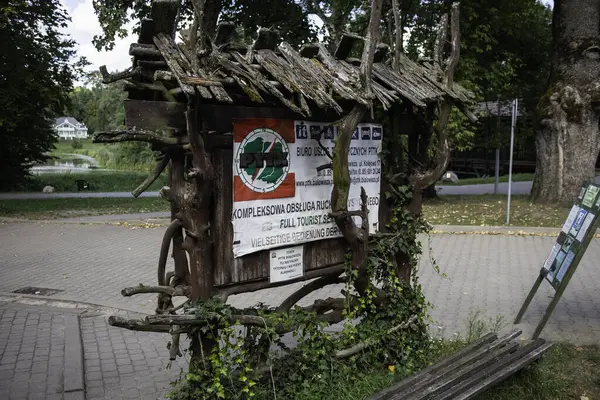 Üzerinde yeşil logo olan bir tabela bir ağaca yaslanıyor. Tabela yosunla kaplı ve ahşap çerçevesi var.