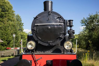 Arka planda ağaçlar olan bir tarlada bir buhar makinesi oturuyor. Tren siyah ve kırmızıdır ve eskidir. Manzara nostaljik ve huzurlu.
