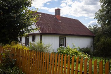 Beyaz çatılı ve kırmızı bacalı küçük bir ev. Ev ahşap bir çitle çevrili ve önünde bir bahçe var.