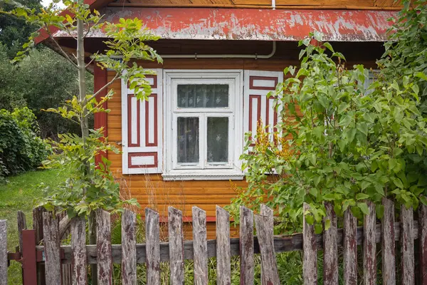 Beyaz pencereli ve ahşap çitli küçük bir ev. Ev ağaçlar ve çalılarla çevrili.