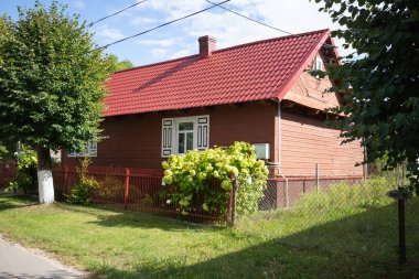 Beyaz çatılı ve beyaz çitli kırmızı bir ev. Ev ağaçlarla çevrili ve küçük bir bahçesi var.