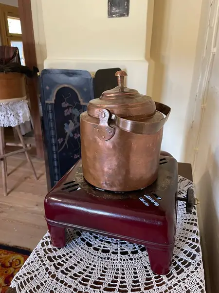 Kırmızı bir standın üzerinde bakır bir tencere duruyor. Eskimiş ve kırsal bir görünüşü var. Stant ahşaptan yapılmış ve üzerinde dantel bir altlık var. Sahne rahat ve sıcak bir atmosfer veriyor.