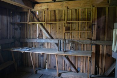 Küçük, boş bir oda, ahşap bir raf ve bir masa. Oda loş bir şekilde aydınlatılmış ve kırsal bir hissi var.