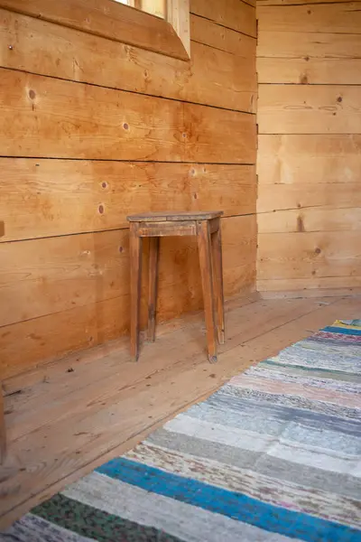 Tahta bir tabure ahşap bir duvarın önünde oturur. Tabure eski ve yıpranmış ve duvar ahşaptan yapılmış. Oda küçük ve rahat, yerde bir halı var.