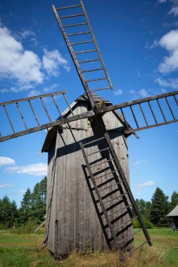 Tahta basamakları olan bir yel değirmeni. Yel değirmeni eski ve kırsal bir görünüme sahip. Gökyüzü açık ve mavi ve arka planda ağaçlar var.