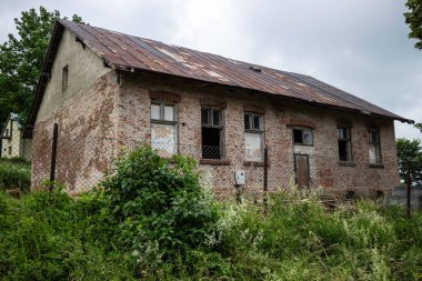 Metal çatılı ve etrafında bir sürü yabani ot büyüyen büyük bir tuğla ev. Ev terk edilmiş ve bakımsız görünüyor.