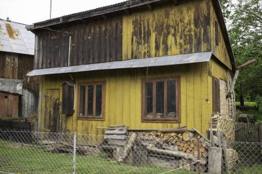 Önünde ahşap bir dış görünüşü ve çiti olan sarı bir ev. Evin önünde yığınla odun ve kütük var.