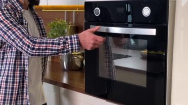 Evde günlük kıyafetlerle yemek pişiren yakışıklı sakallı adam. Pozitif ev erkeği fırında kavrulmuş yemekleri kontrol ediyor, sebze kokusu alıyor, ev mutfağında yemek hazırlıyor. Yakın görünüm