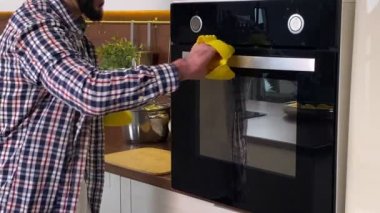 Evde günlük kıyafetlerle yemek pişiren pozitif sakallı bir adam. Fırın eldiveni takan neşeli bir adam, sobayı açıyor, tavayı kızarmış yiyeceklerle alıyor, içerideki kokuyu tatmin ediyor. Ev erkeği yemek hazırlıyor.