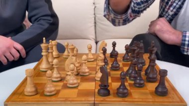 Satranç oynayan iki adamın elleri kapalı alanda siyah beyaz tahta parçalarla satranç tahtasına hamle yapıyor. Arkadaşlar mantıksal masa oyununda hareket üzerine kafa yorar. Ev eğlencesi, hobi eğlencesi.