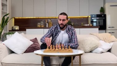 Orta yaşlı bir adam evinde kanepesinde oturarak satranç oynuyor. Sakallı adam mantıklı bir masa oyununda hareket eder. Beyaz siyah satranç taşları, şah mat. Ev eğlencesi, hobi eğlencesi.