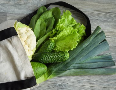 Composition of green vegetables in eco fabric bag on wooden table. Top table view. Pile of cucumber, avocado, lettuce, leek, spinach, cauliflower in reusable shopping bag. Grocery shopping concept
