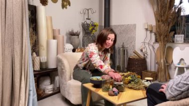 Skilled female herbalist mixing various dried herbs with mortar pestle while holding appointment male client indoors. Middle-aged woman using natural herbal methods of treatment. Alternative medicine