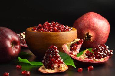 Wooden mortar full ripe arils of pomegranate, red apples, green leaves on black background. Still life fruits, sweet seeds. Split open clusters of pomegranates. Healthy food. Studio shot