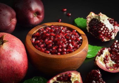 Wooden mortar full ripe arils of pomegranate, red apples, green leaves on black background. Still life fruits, sweet seeds. Split open clusters of pomegranates. Healthy food. Studio shot