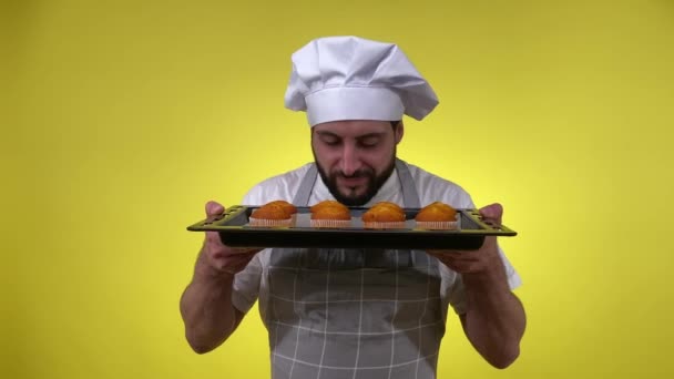 头戴围裙 头戴帽子 满脸喜气的大胡子男人在做甜点 拿着烤盘 散发着淡淡的淡淡淡的香味和淡淡的黄色背景 快乐的男性厨师在烤箱里放着美味的松饼 — 图库视频影像