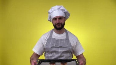 Önlük şapkalı, hayal kırıklığına uğramış orta yaşlı erkek aşçı kek pişirmede başarısız, sarı arka planda izole edilmiş fırın tepsisindeki yanmış kekleri gösteriyor. Üzgün aşçı içeride tatlıyı fazla pişiriyor. Mutfak konsepti