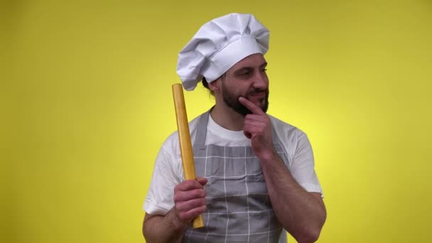 头戴围裙头戴蓬乱胡须的男人想出菜谱 准备烹调晚餐 拿着木制滚针隔离黄色背景 快乐的男厨师有好吃的菜的主意 烹饪概念 — 图库视频影像