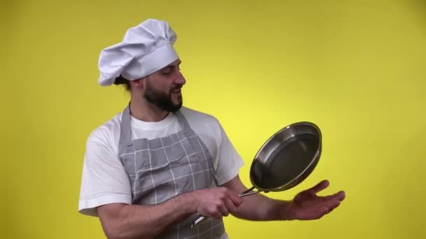 黄色の背景に隔離されたフライパンを保持している陽気な男性料理人 調理の準備ができてエプロンの帽子で中年の男性を笑顔 夕食を作るための台所用品を準備します 料理の概念 — ストック動画