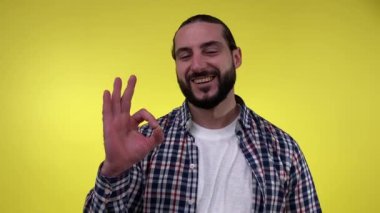 Resimli, sakallı, gülümseyen, işaret parmaklarıyla gülümseyen, sarı arka planda izole edilmiş günlük gömlekler giyen adam. Erkek insan jest yapıyor, tamam diyor, her şey yolunda. Başarılı ifade