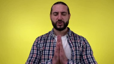 Sakallı adamın portresi dua ediyor, yalvarıyor, el ele tutuşuyor ve sarı arka planda izole edilmiş umut dolu bir yüz ifadesi var. Tişört giyen, af dileyen, gözyaşı döken endişeli bir erkek.