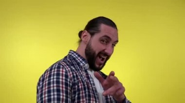 Oyuncak erkek giyimli, günlük kıyafetlerle flört eden, içerde kameraya parmak tabancalarını gösteren. Resimli sakallı adam kamerayı işaret ediyor, göz kırpıyor, sarı arka planda izole edilmiş kameraya bakıyor..