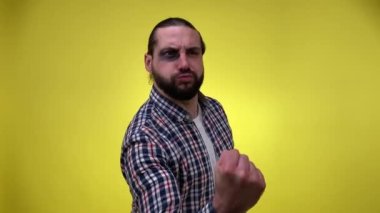 Gözü morarmış, yumruğunu sallayan, tehdit eden orta yaşlı bir adam. Sakallı bir erkek kazadan sonra göz yaralanması geçirir ya da morluklarla dövüşür. Sarı arka planda izole edilmiş kapalı stüdyo görüntüleri.