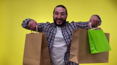 Orta yaşlı adam alışverişinden memnun, alışveriş merkezinden alınmış alışveriş torbalarını gösteren yeni alışverişleri takdir eden, sarı arka planda izole edilmiş paketler tutan. Tüketim kavramı