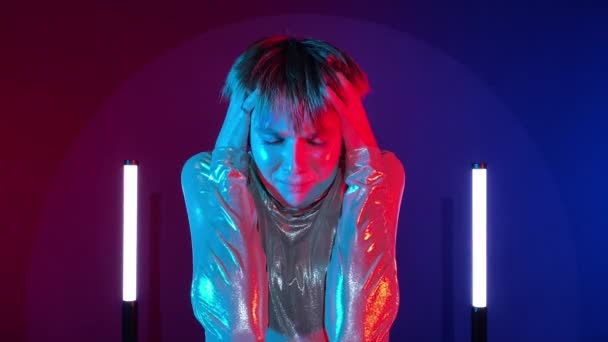 女机器人穿着未来派服装 头上有噪音 耳朵上有手 有心理干扰 在室内有霓虹灯照明时精神控制 年轻的成年女子在艺术五彩缤纷的照明 Sci — 图库视频影像