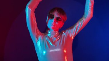 Genç, yetişkin bir kadın, kapalı alanda renkli ışıkta dans ediyor. Yuvarlak güneş gözlüğü takan parlak elbiseli, kısa saçlı bir kadın. Gece hayatı, gece kulübü konsepti.