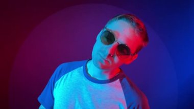 Kapalı alanda renkli ışıkta dans eden dikkatsiz yüz ifadesi olan genç yetişkin bir adam. Yuvarlak güneş gözlüğü takan, kısa saçlı bir erkek. Gece hayatı, gece kulübü konsepti.