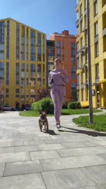 Mor spor giyim giymiş genç bir kadın, güneşli bir sabah, şehirde köpek yürüyüşü yapıyor. Dışarıda evcil hayvan besleyen sağlıklı bir evcil hayvan sahibi. Şehir yaşam tarzı konsepti