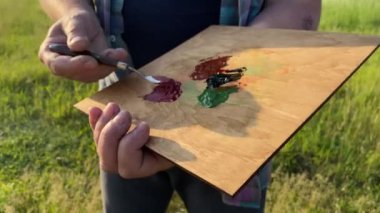 Tarlada çalışan model sakallı bir ressam, palet bıçağıyla gün doğarken açık havada yağlı boya kullanarak resim yapıyor. Ahşap sehpada sanatçı tuvali, malzeme malzemeleri boyama. Manzara görünümü, doğa arkaplanı