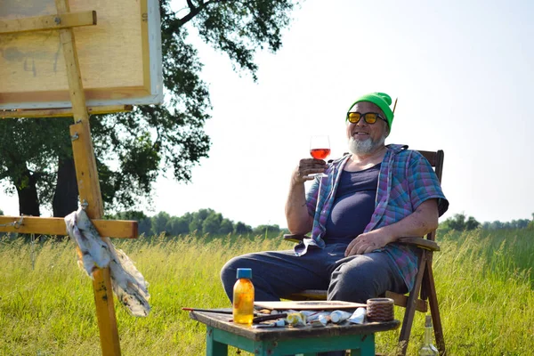 フィールドワーク終了後にリラックスした大人の芸術家をオフビート 朝の日差しの中で屋外の椅子に座って絵を見てワインを飲みます 木製のイーゼル塗装ツールのキャンバス材料供給 — ストック写真