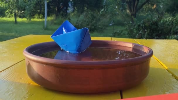 平衡概念 蓝色纸船在粘土碗中的水面上航行 五颜六色的折纸雕像躺在外面的黄色桌子上 背景上的自然景观 孤独的想法 — 图库视频影像