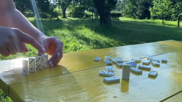 平衡概念 难以辨认的女人在室外阳光下把一排多米诺骨牌放在黄色的桌子上 背景是美丽的园林 女性的手平衡着多米诺骨牌 一个接一个的放下来 — 图库视频影像