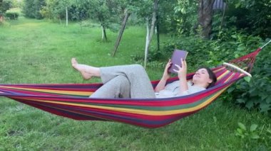 Neşeli yetişkin kadın, güneş doğarken yeşil manzara parkı bahçesindeki renkli hamakta dinlenirken kitap okuyor. Hayat boyu denge işi konsepti. Doğada boş vakti olan bir kadın, izin günü. Rekreasyon