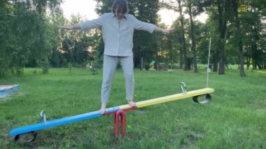 Sevimli yetişkin kadın gün batımında tahterevallide dengede duruyor. Denge konsepti. Çıplak ayaklı kadın çocuk parkı ekipmanında denge buluyor. Hayat iş dengesi. Arkaplanda yeşil peyzaj