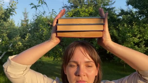 年轻的成年女子在日落时闭着眼在头上平衡书本 平衡概念 生活工作平衡类比 金光闪闪的阳光映衬下的绿色园林公园 — 图库视频影像
