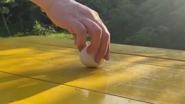 在外面的黄桌子上旋转着白蛋 平衡概念 女性的手旋转物体作为平衡生活的类比 金色的阳光照射在表面 背景上的绿色花园 — 图库视频影像