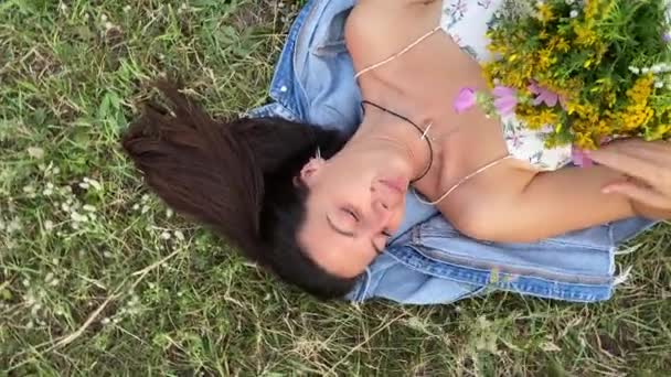 美丽的年轻女子在大自然的美丽中享受着轻松的消遣 她躺在草地上 捧着五颜六色的野花束 闻着难闻的野草味 直接在旋转弹丸上方 — 图库视频影像