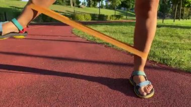 Çok ırklı genç bir kadın sabah güneşinde halka açık parkta egzersiz yapıyor. Direnç bandını kullanarak bacak esnetme hareketleri yapıyor esnekliği arttırıyor, şafakta güç kazanıyor. Etkin yaşam biçimi